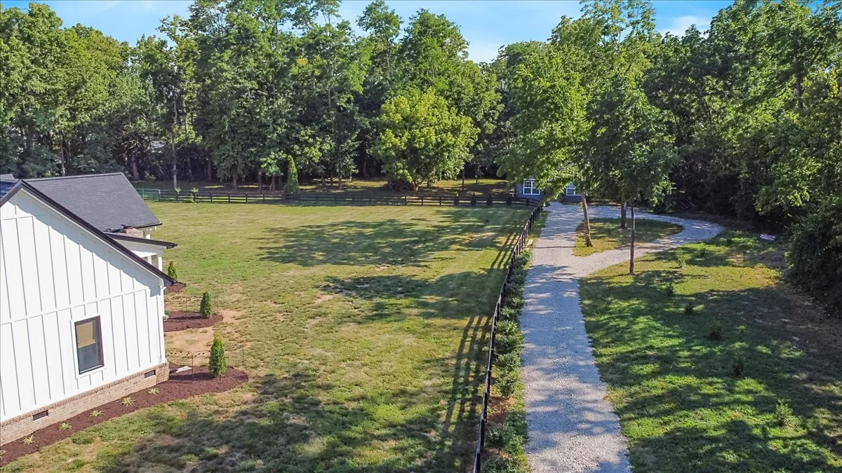 Aerial view of a property with a white house, a gravel walkway, green grass, trees, and a wooded background.