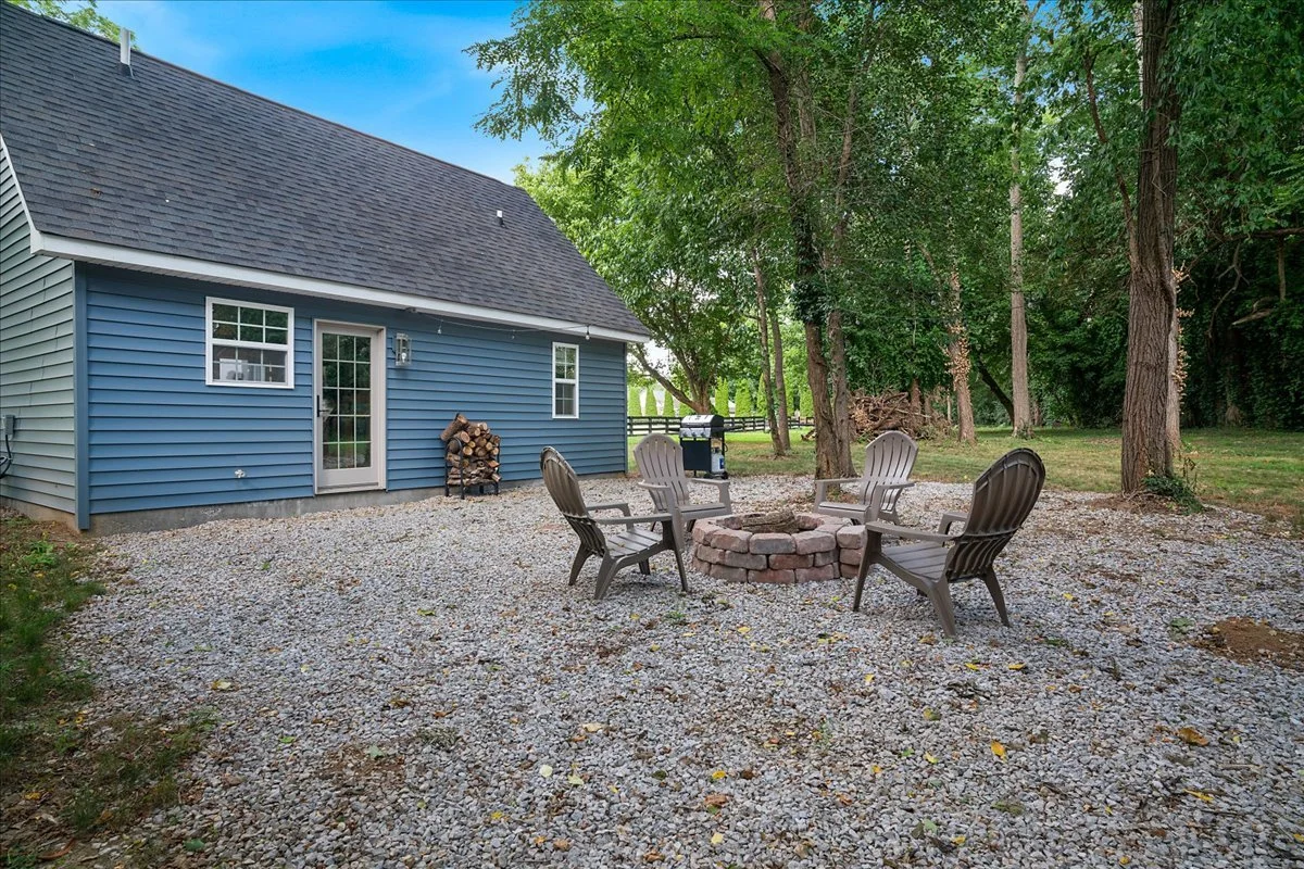 Backyard with gravel patio, round brick fire pit surrounded by four Adirondack chairs, trees providing shade, house with blue siding and a glass door, woodpile, and a barbecue grill.