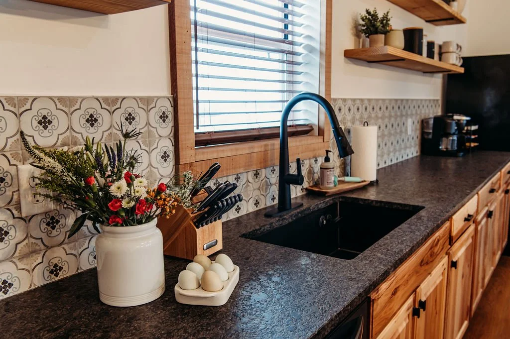 Kitchen countertop with a vase of flowers, a knife block, a roll of paper towels, soap dispenser, and a black sink with a black faucet. Wooden shelves with plants, books, and mugs on the wall, window with blinds, and a coffee maker in the background.