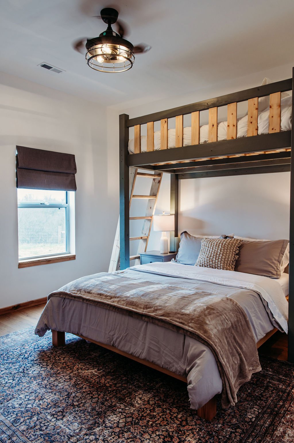 A bedroom with a bed, a lofted area with a ladder, a window with a brown shade, a nightstand with a lamp, and a ceiling fan in a modern rustic style.