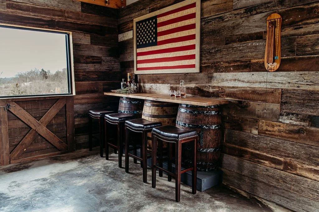 A rustic indoor bar area with a wooden table and four bar stools, a large American flag on the wall, and a bottle of liquor. The room has wood-paneled walls and a window showing a outdoor landscape.