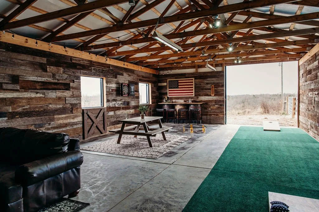 An indoor rustic space with wood plank walls, exposed wooden ceiling beams, a concrete floor with a section of green turf, a black leather couch, a wooden picnic table with a plant, a dartboard, an American flag, a wooden bar with stools, and a large