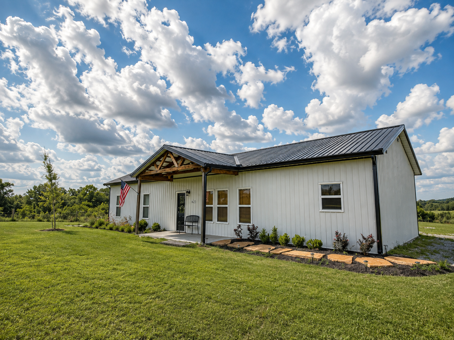 White house with a metal roof, front porch with black bench, American flag, and landscaped garden, under a partly cloudy sky.