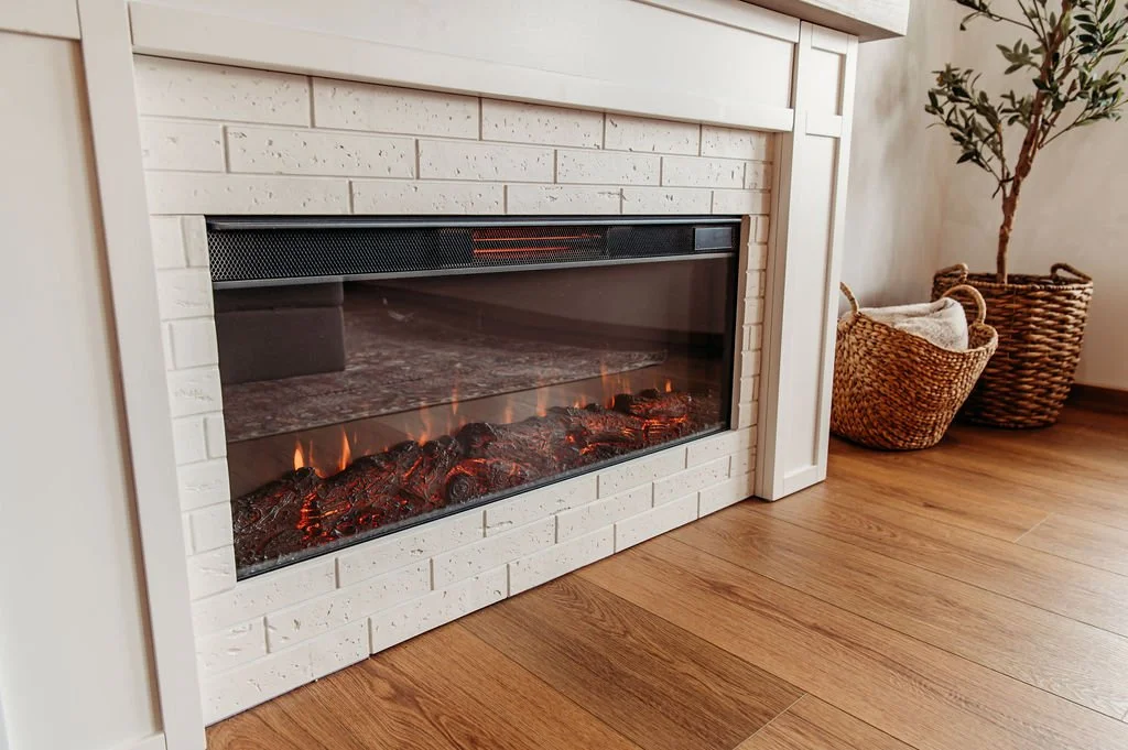 Electric fireplace with faux logs inside a white brick surround, next to woven baskets with textiles and a potted plant on hardwood floor.