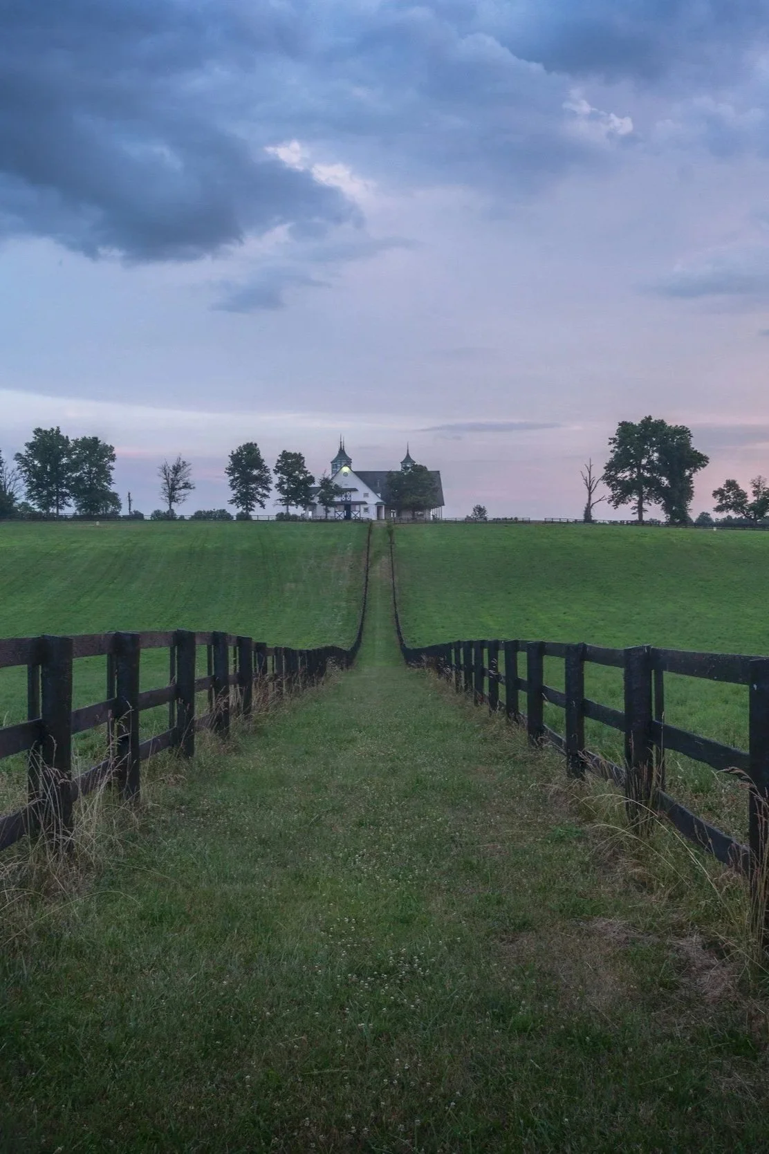A grassy path flanked by black fences leading up a gentle hill to a white church with two steeples, set against a cloudy sky at dusk.
