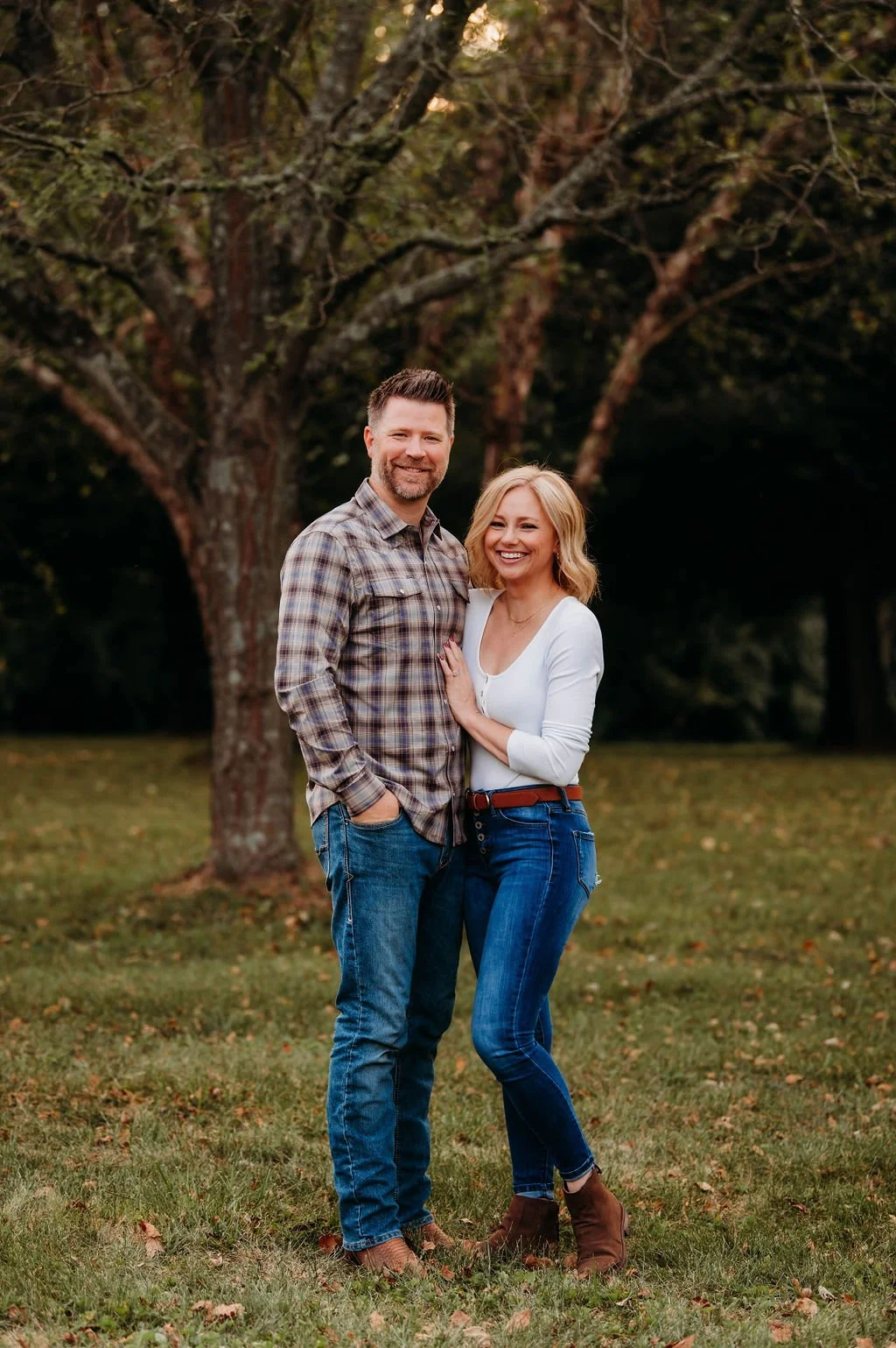 A smiling man and woman stand close together outdoors, with a large tree and green grass in the background. The man has short dark hair and a beard, wearing a plaid shirt and jeans. The woman has shoulder-length blonde hair, wearing a white top, blue jeans, and brown boots. They are happy and appear to be enjoying a moment together.