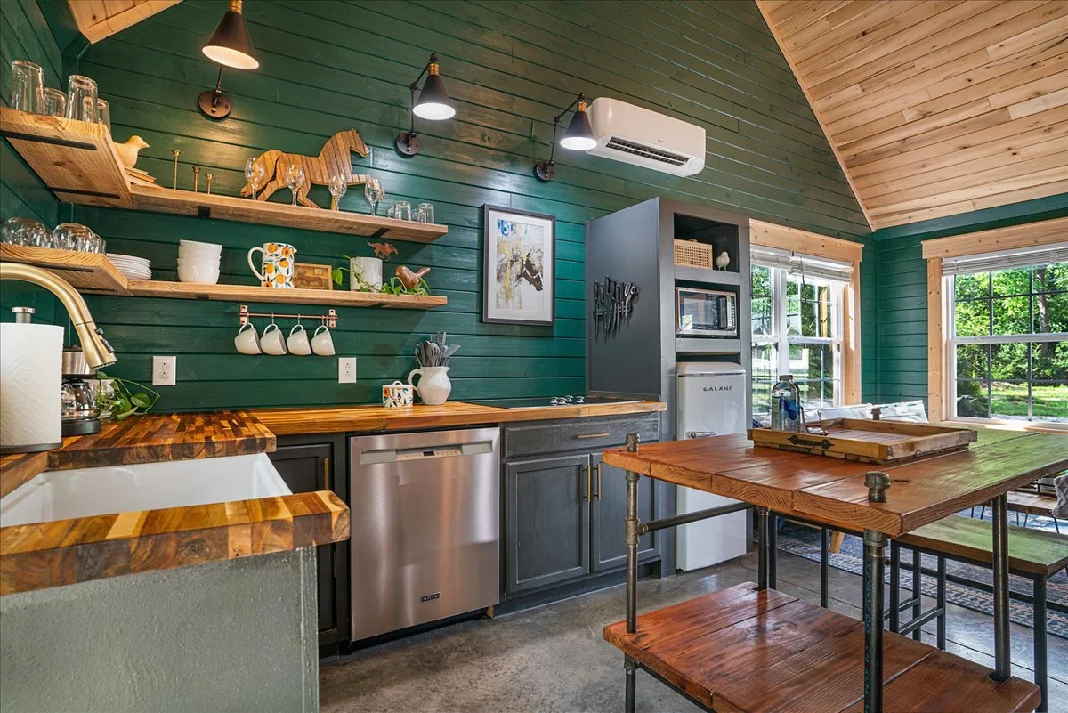 Kitchen with wooden accents, green paneled walls, open shelves, stainless steel appliances, and natural sunlight coming through large windows.