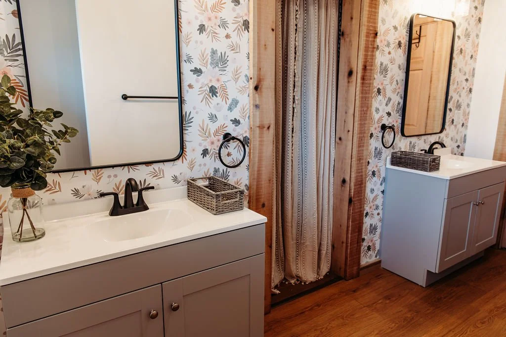 A bathroom with two gray vanities with white countertops, each with a mirror above them. The left vanity has a plant in a glass vase, and the right vanity has a basket. A wooden beam separates the two vanities, with a patterned curtain hanging from i
