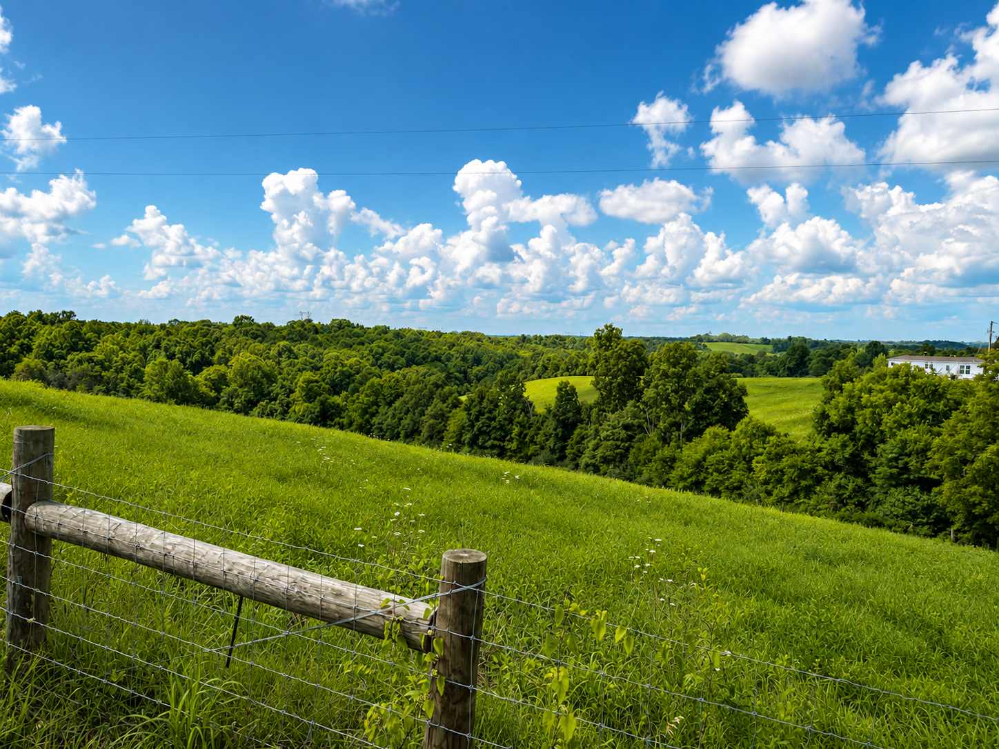 Green rolling hills, dense trees, and a blue sky with fluffy white clouds in a rural landscape with a wooden and wire fence in the foreground.