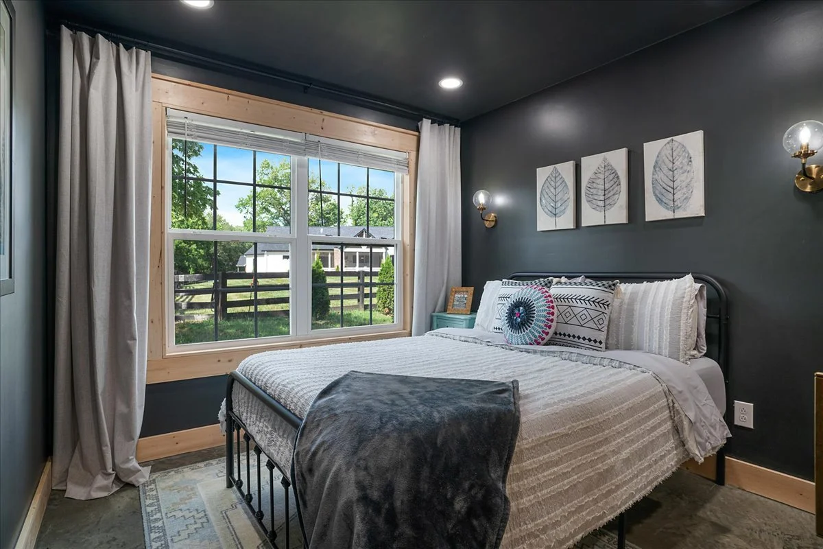 A cozy bedroom with a large window showing a view of green trees and a white house outside; dark gray walls with three framed leaf prints above the bed; bed with white and patterned pillows and a gray blanket at the foot; brass wall sconces on either