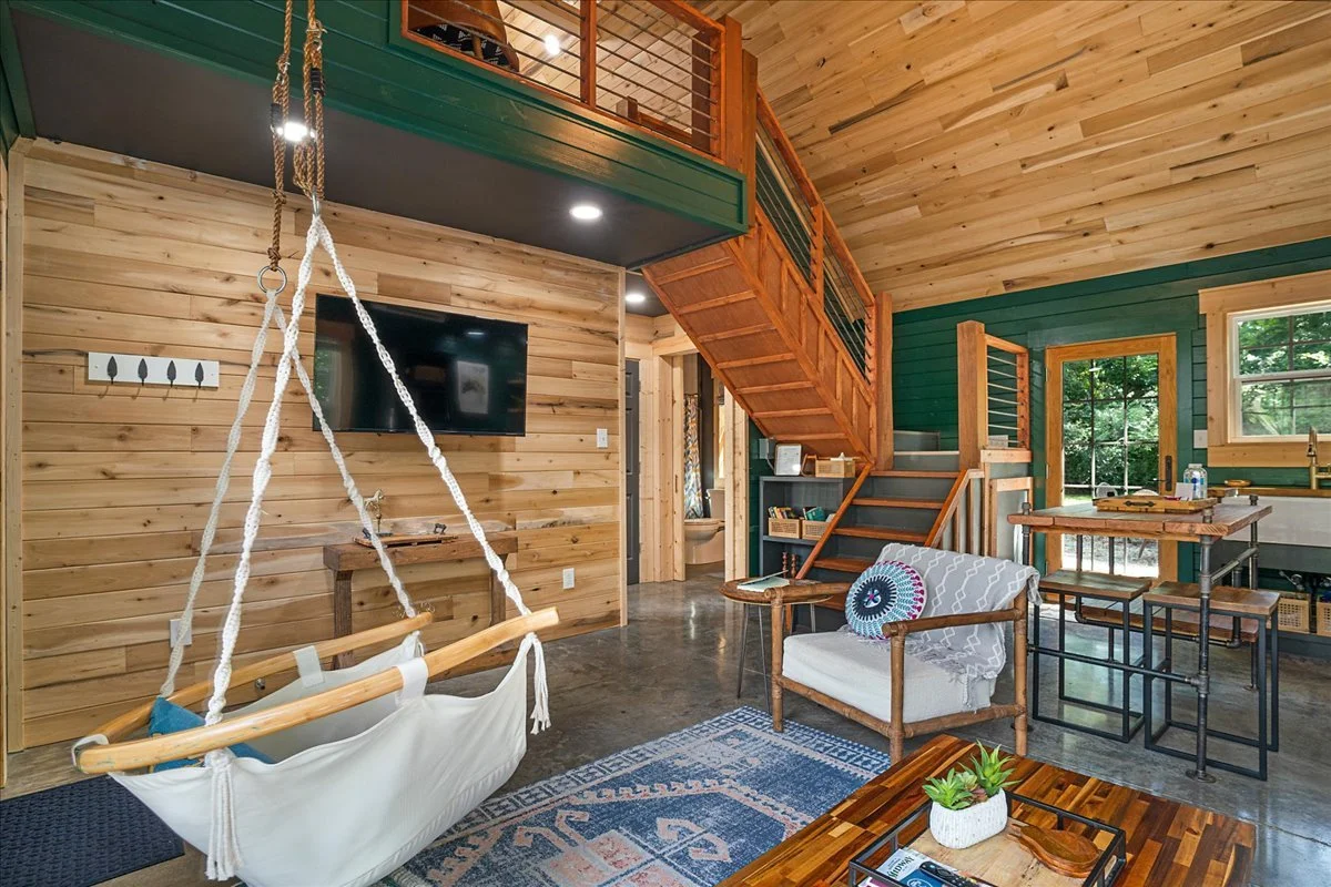 A cozy living space with wood-paneled walls and ceiling, featuring a hanging swing, a flat-screen TV, a staircase, and a sitting area with a chair, coffee table, and rug, all illuminated by natural light from large windows.