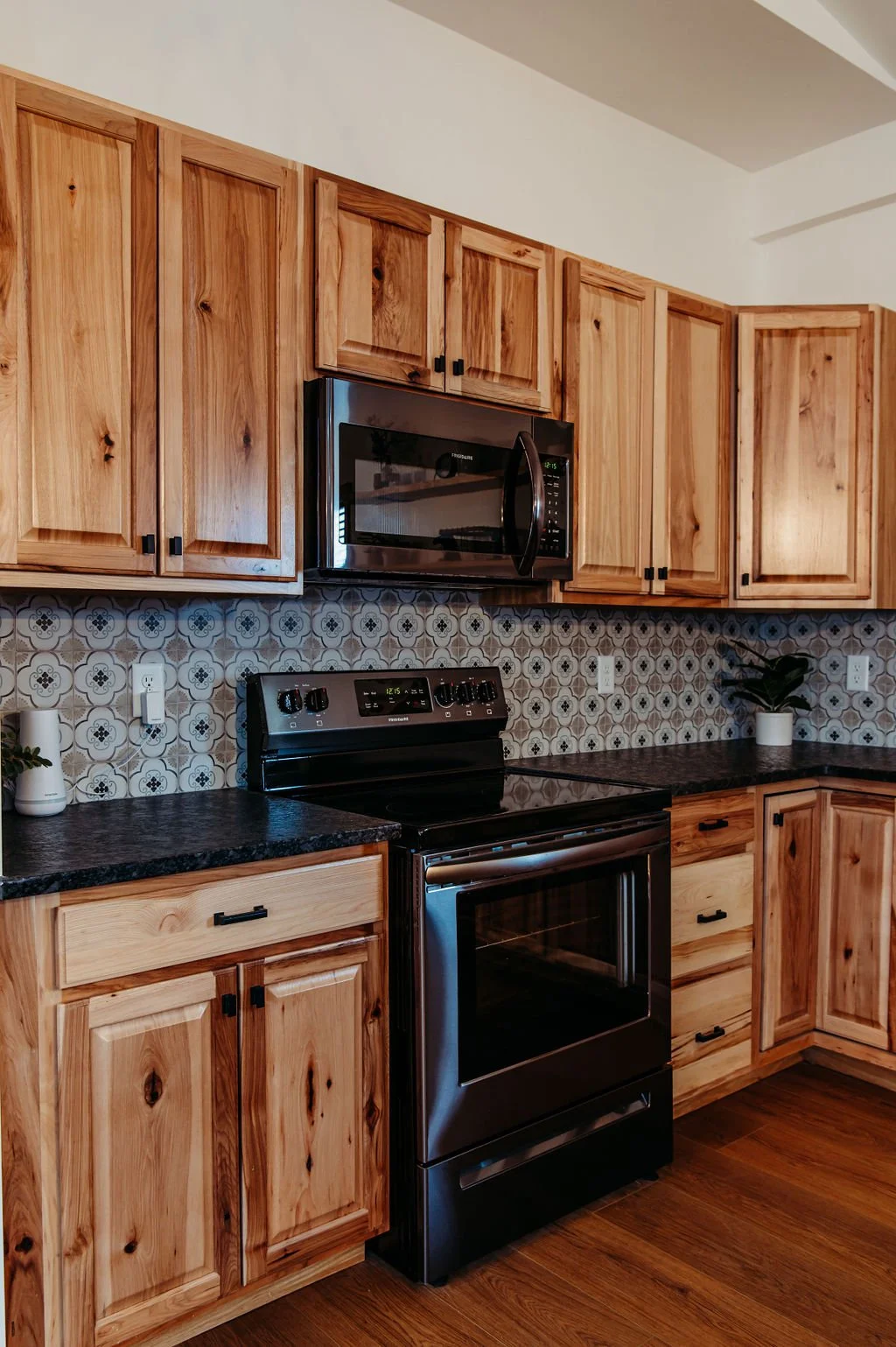KITCHEN with wooden cabinets, black microwave, black stove, patterned tile backsplash, and potted plants.