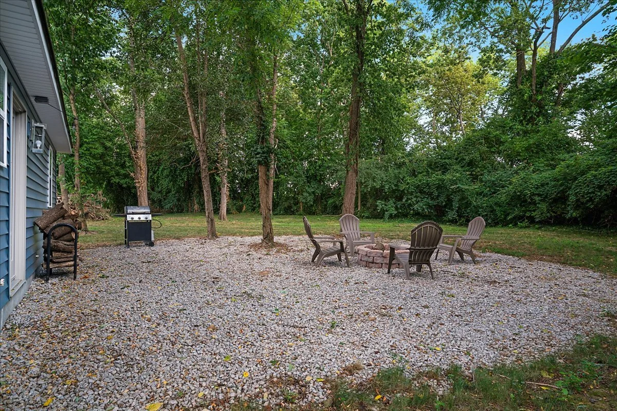 Backyard patio area with gravel ground, four Adirondack chairs arranged in a circle around a fire pit, picnic table, and grill, surrounded by trees and greenery.