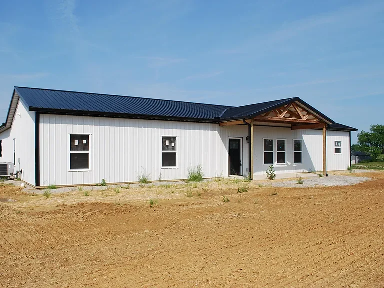 Newly constructed single-story house with white siding, black metal roof, and a covered porch with wooden support beams, situated on a dirt lot.