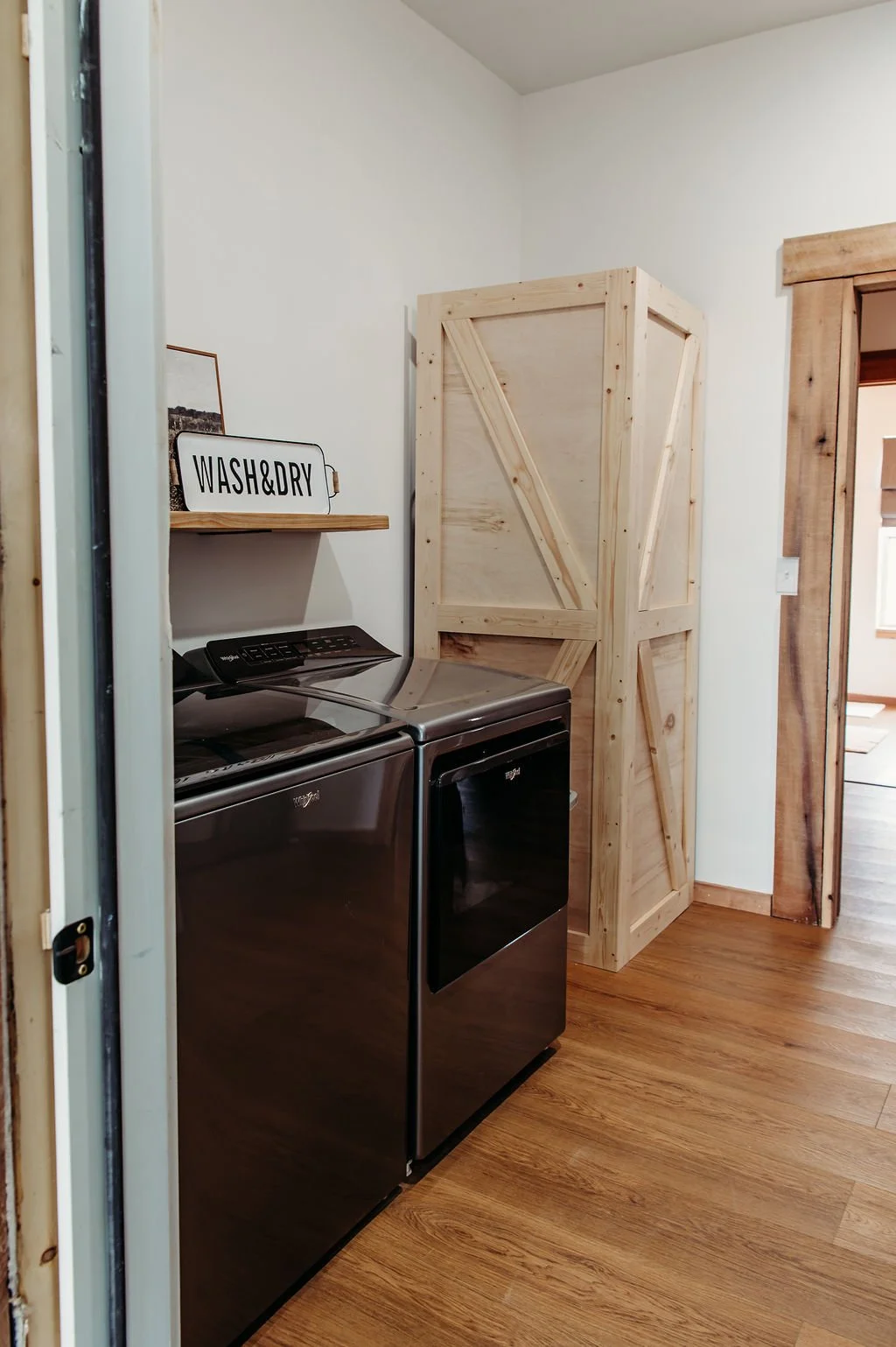 Laundry room with a black washer and dryer, a wooden shelf with a wash & dry sign, and a wooden storage cabinet, wood flooring, and a doorway.