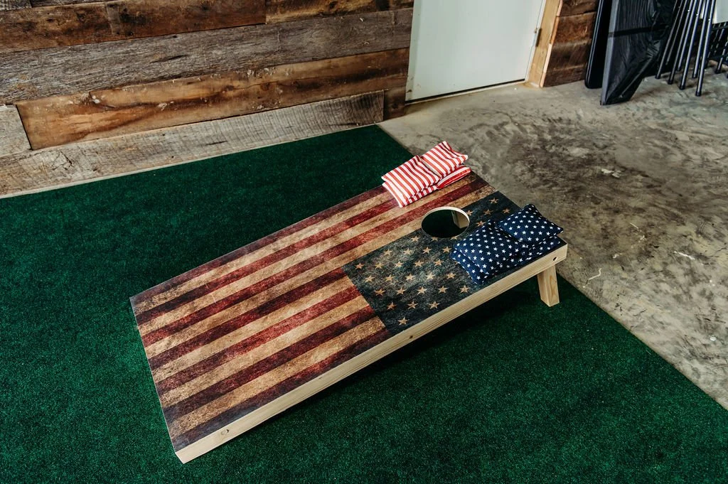 A wooden cornhole board decorated with an American flag design, positioned on green turf. Two small pillows with patriotic patterns and a round black object are placed on the board.