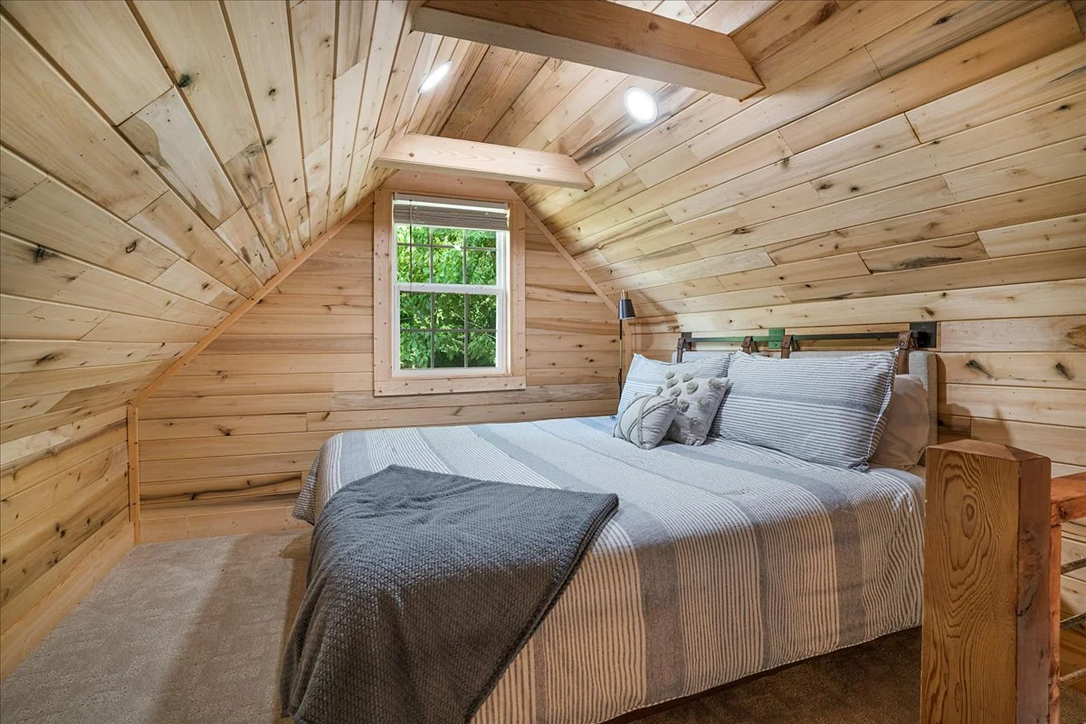 A bedroom with wood-paneled sloped ceiling, a window showing trees outside, a bed with striped bedding and decorative pillows, and a wooden nightstand.