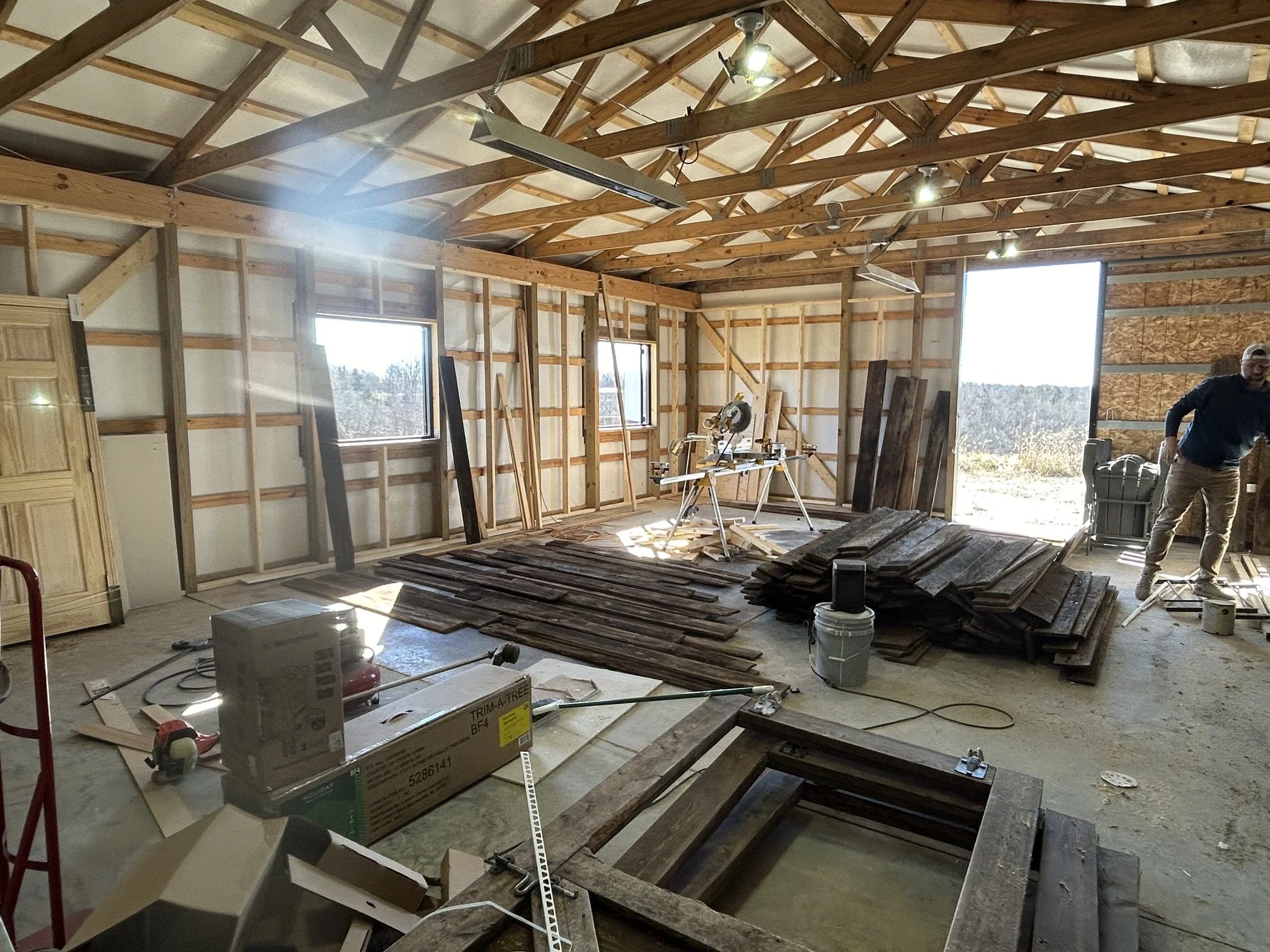 Construction inside a partially built wooden structure, with framed walls and a high ceiling, a man in work clothes standing near an open doorway, construction tools and materials scattered around, including stacks of wood, a circular saw, and a yellow measuring tape.