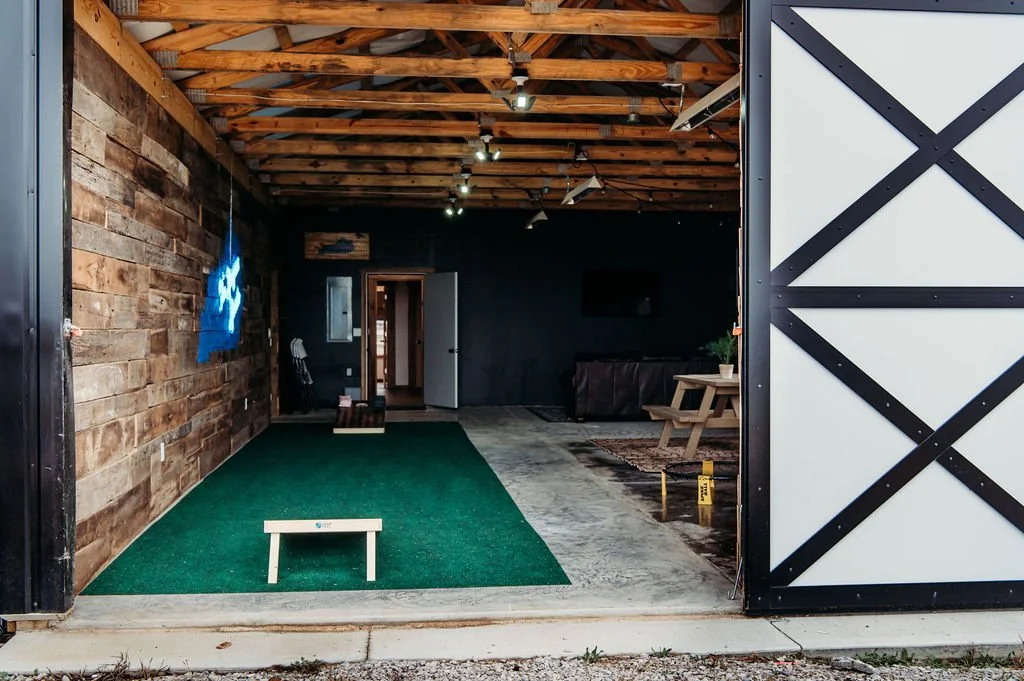 Indoor golf hitting bay with artificial turf, wooden walls, and ceiling beams, a small bench, and a seating area with a table and a TV.