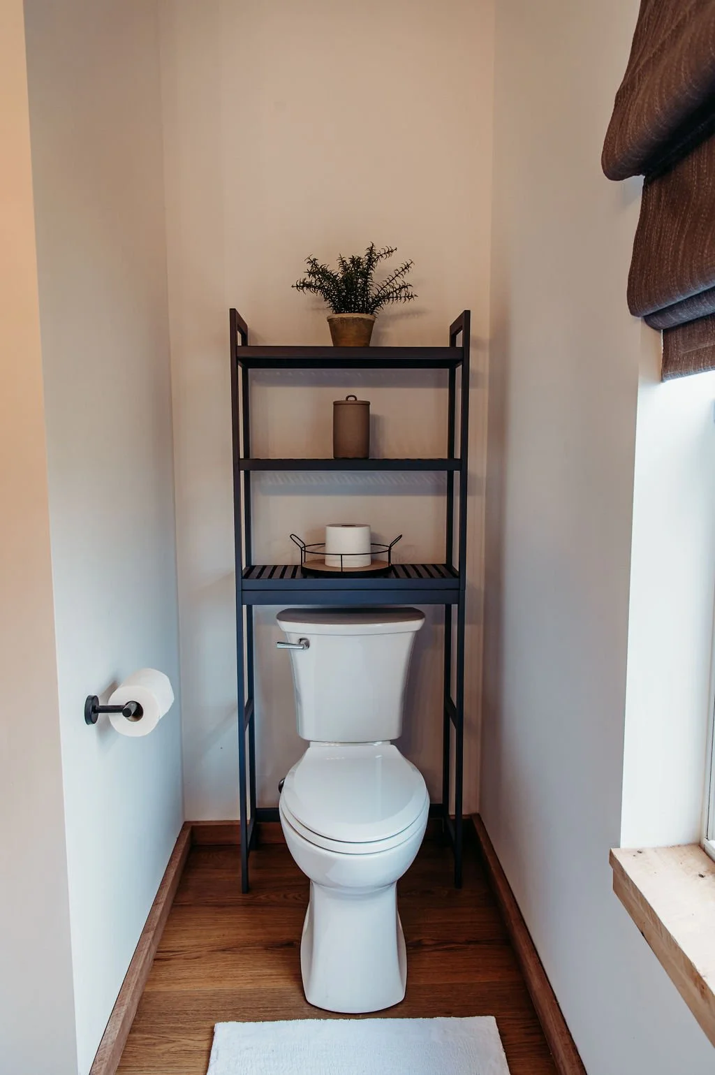 Bathroom with a white toilet underneath a black metal shelving unit. The top shelf has a potted plant, the middle shelf has a brown container, and the lower shelf has a paper towel roll on a tray. A white rug is placed in front of the toilet, and a w