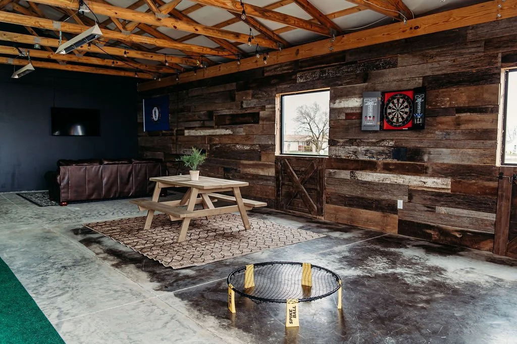 An indoor recreational space with a wooden ceiling, a dark accent wall, and a rustic wood-paneled wall. It features a dartboard, a flat-screen TV, a brown leather couch, a wooden picnic table with a potted plant, an area rug, and a small yellow tramp