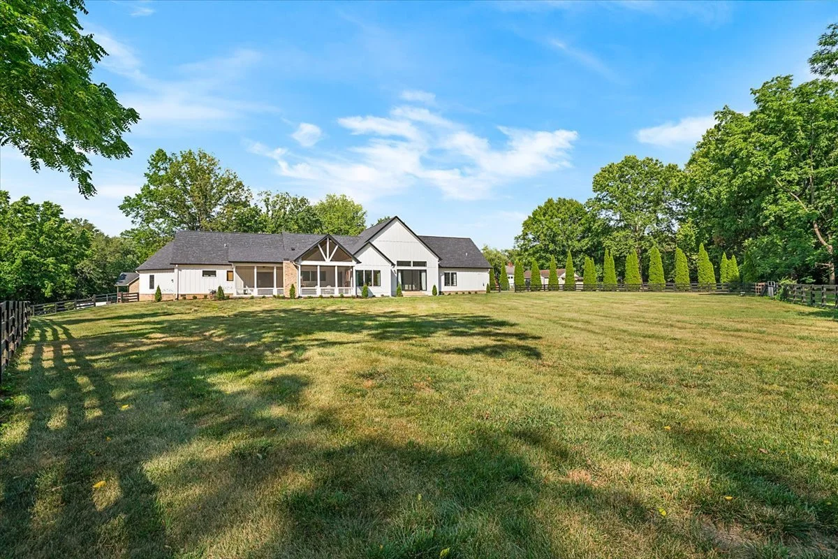 A large white house with black roof and trim, surrounded by a vast green lawn with shadows, tall trees, and a blue sky with some clouds.