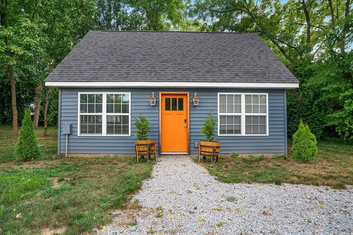 Small house with blue siding, orange front door, gravel pathway, two potted trees, and white window shutters surrounded by green trees.