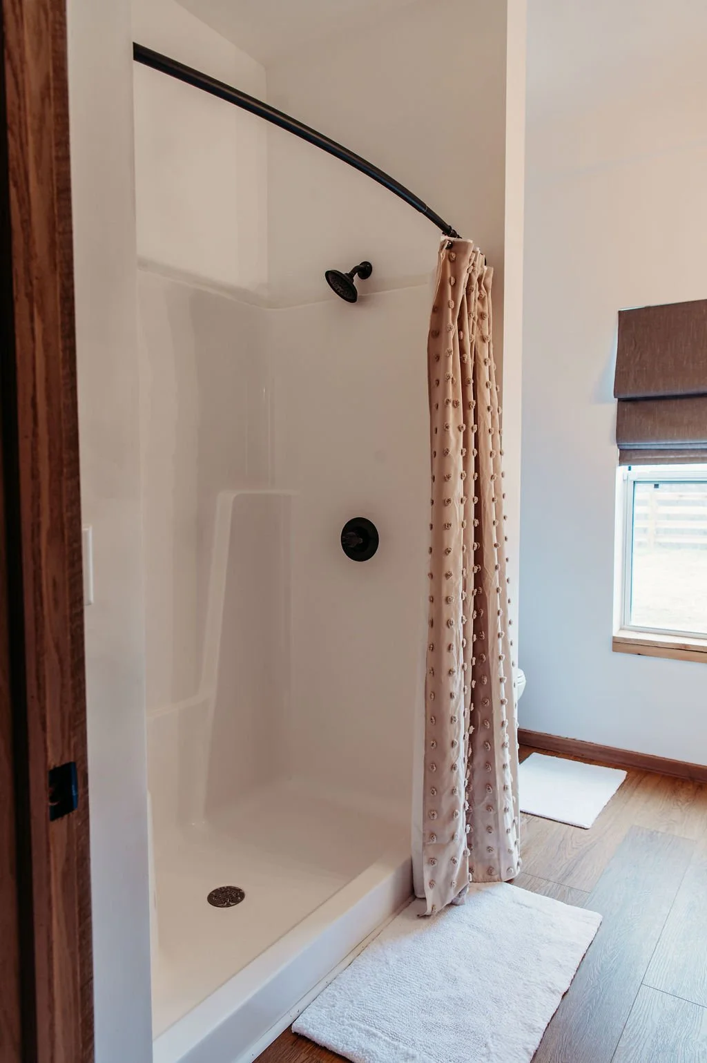 Bathroom shower with a curved shower curtain rod, beige shower curtain, white shower stall, wooden floor, window with a brown shade, and white bathroom floor mats.