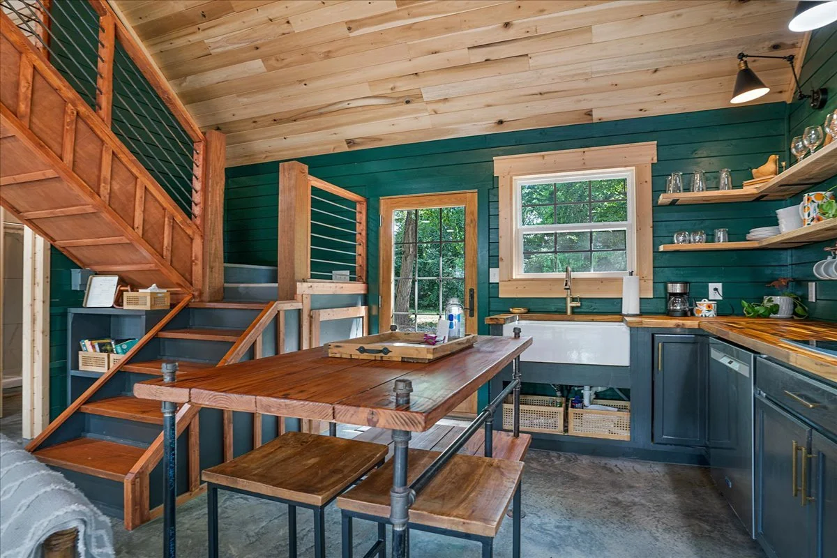 Rustic kitchen with dark teal cabinets, wooden shelves, and a large farmhouse sink, featuring a dining table with matching benches.