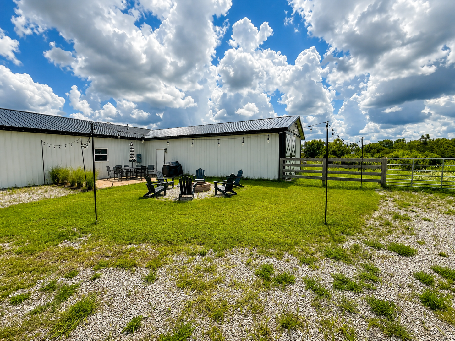 A backyard with a grassy area, a fire pit surrounded by chairs, a gravel pathway, a white barn with string lights, outdoor furniture, and a partly cloudy sky.