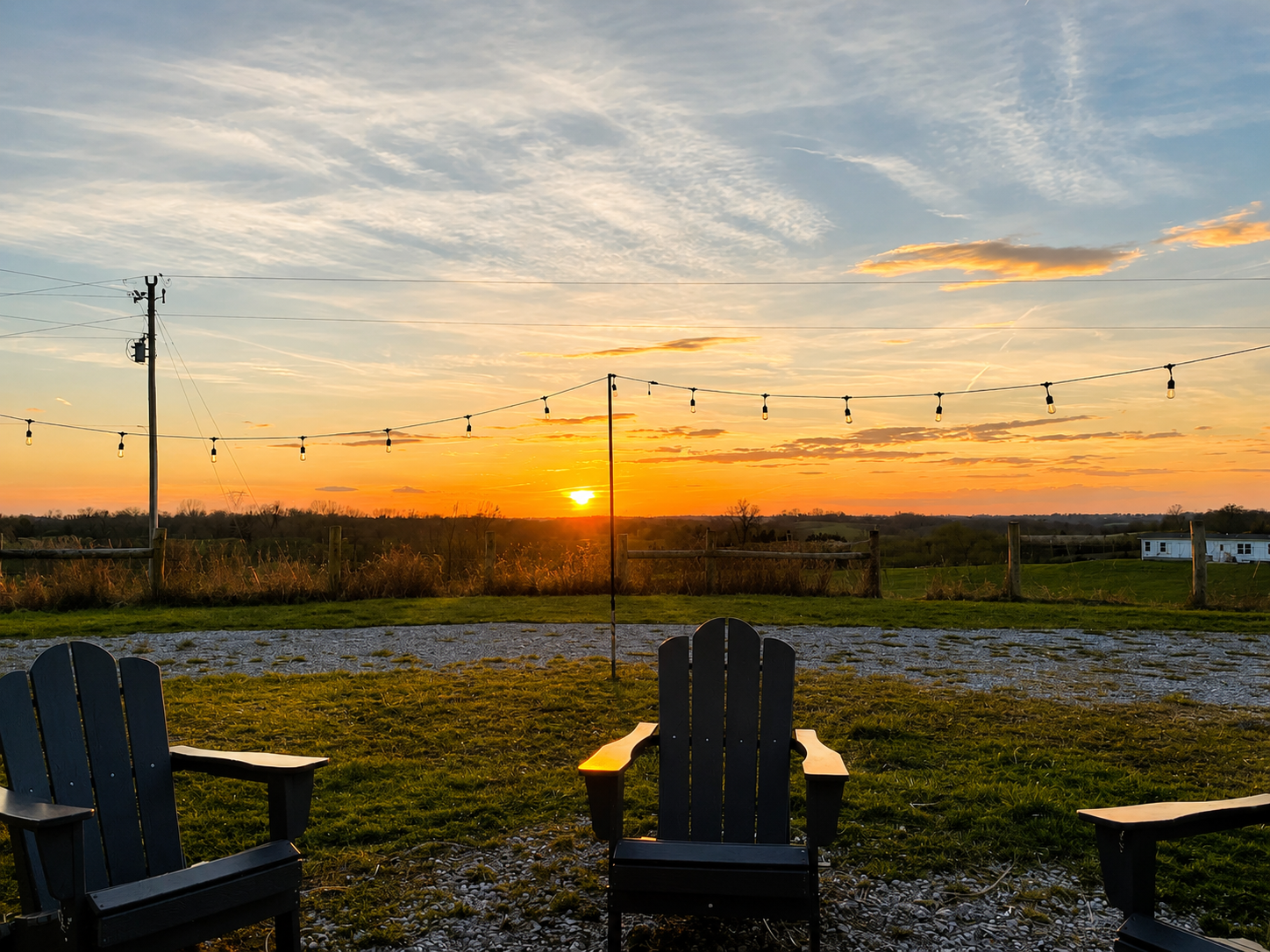 Sunset over a rural landscape with string lights and Adirondack chairs on a grassy area