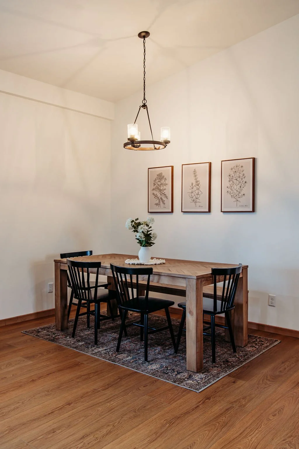 A dining room with a wooden table, four black chairs, a white vase with white flowers, framed botanical art on the wall, a chandelier with two lights, and hardwood floors with a patterned rug.