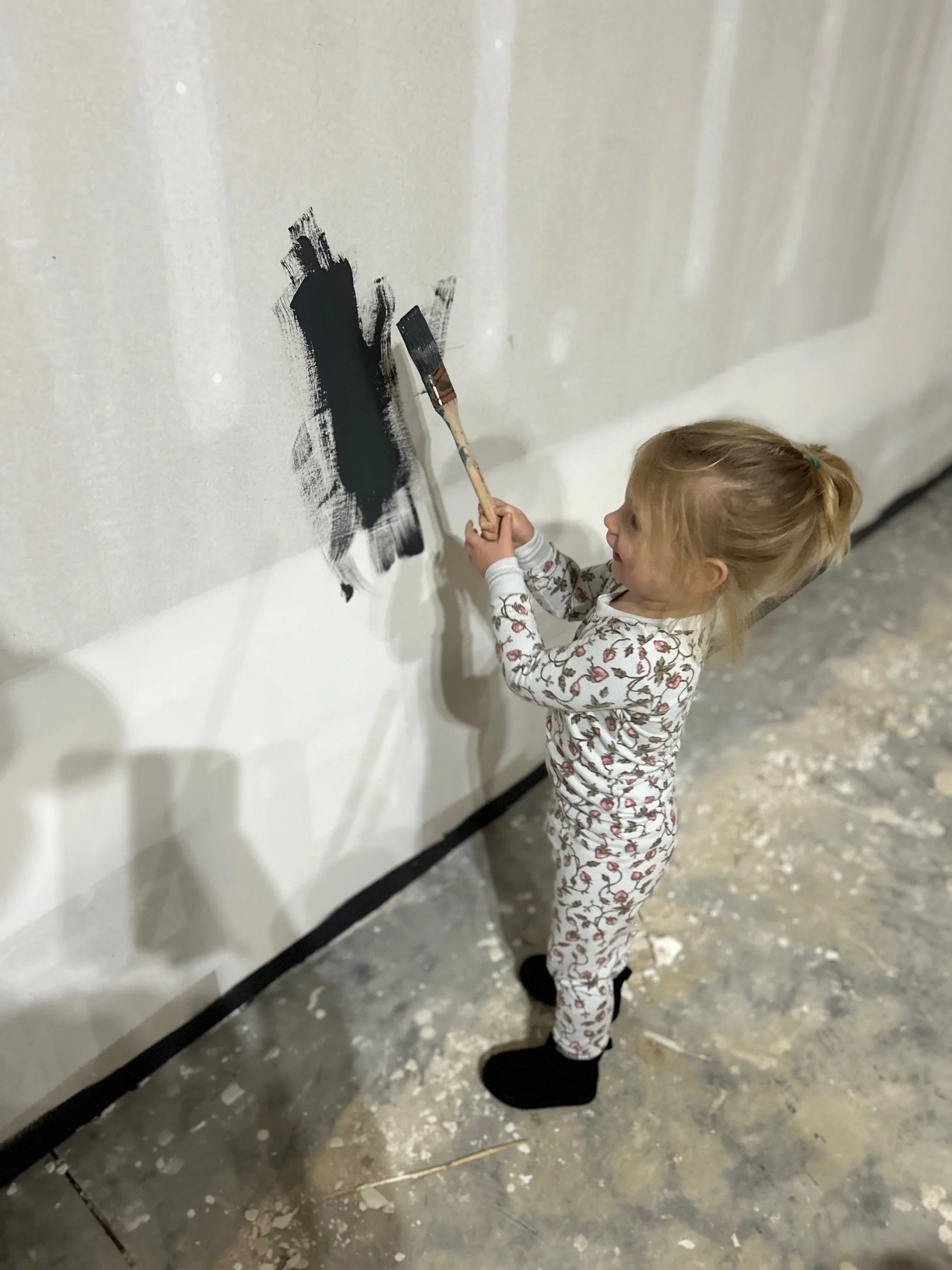 A young girl with blonde hair in pajamas with a flamingo pattern paints black paint on a wall using a small paintbrush.