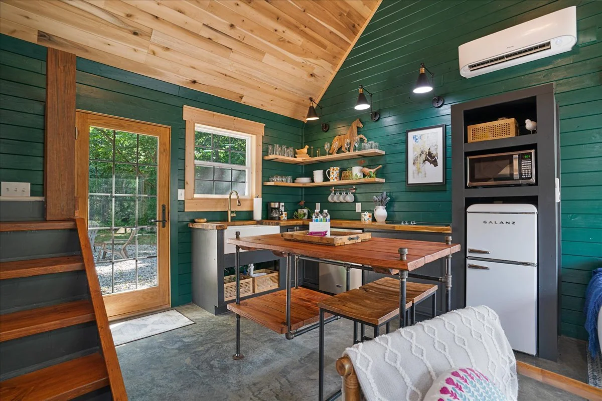 Cozy kitchen with green paneled walls, wooden ceiling, and a sliding glass door leading outside. Contains a small island with a wooden top, open shelves with dishes and decorative items, a coin-op fridge, microwave, and a wall-mounted air conditioner