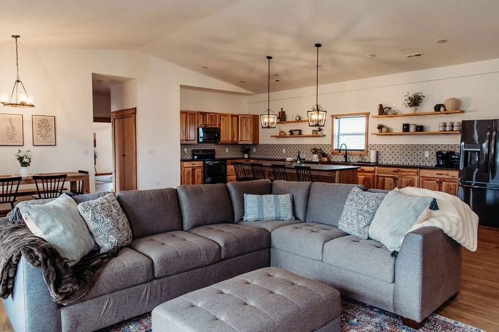Open-concept living room and kitchen with gray sectional sofa, wooden kitchen cabinets, open shelves, patterned backsplash, and black refrigerator.