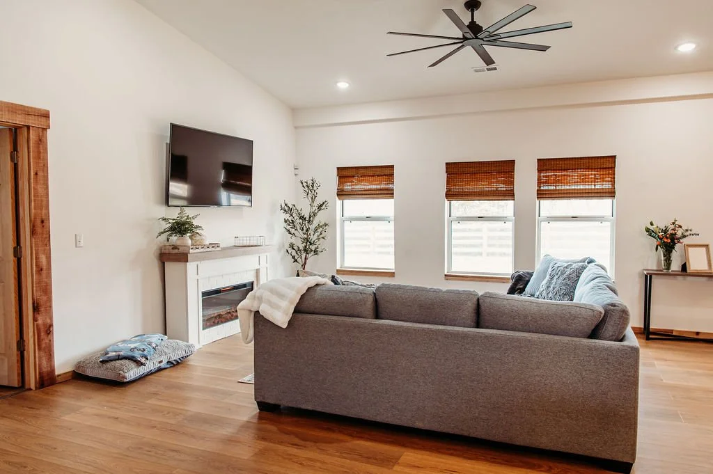 Living room with gray sofa, wooden floors, three windows with brown blinds, a wall-mounted TV above a white fireplace, a side table with flowers, and a modern ceiling fan.