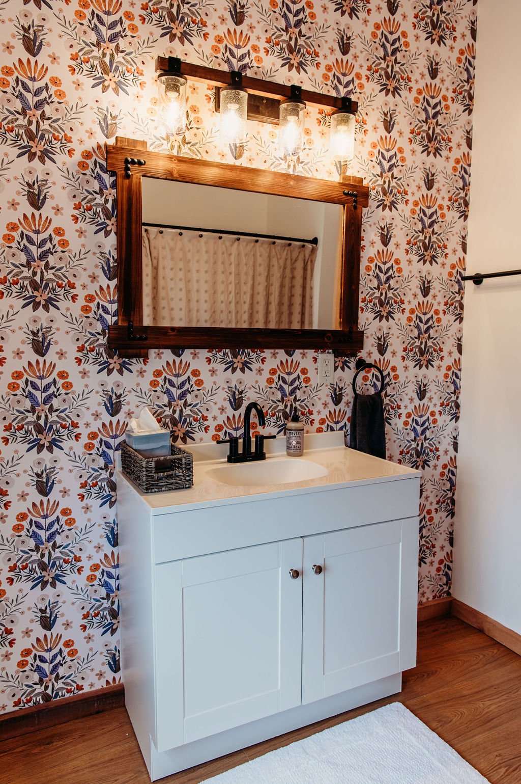 Bathroom with a floral-patterned wallpaper, a white vanity with a black faucet, a mirror with a wooden frame, and overhead lighting fixtures.