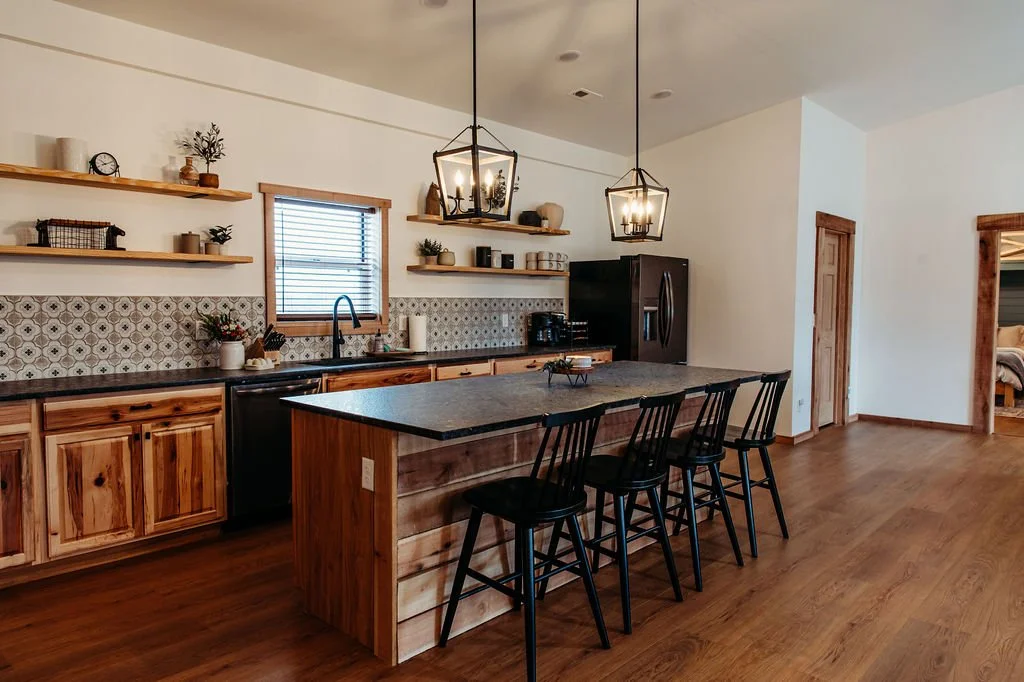 Kitchen with wooden cabinets, a kitchen island with four black chairs, open shelves, and two pendant lights hanging over the island.