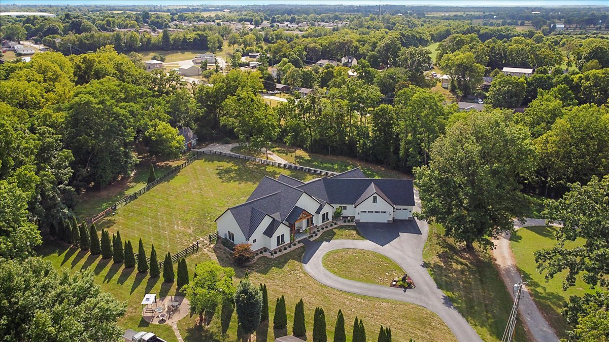 Aerial view of a large white house with dark roof, surrounded by lush green trees and landscaped yard with circular driveway, fencing, and outdoor seating area.