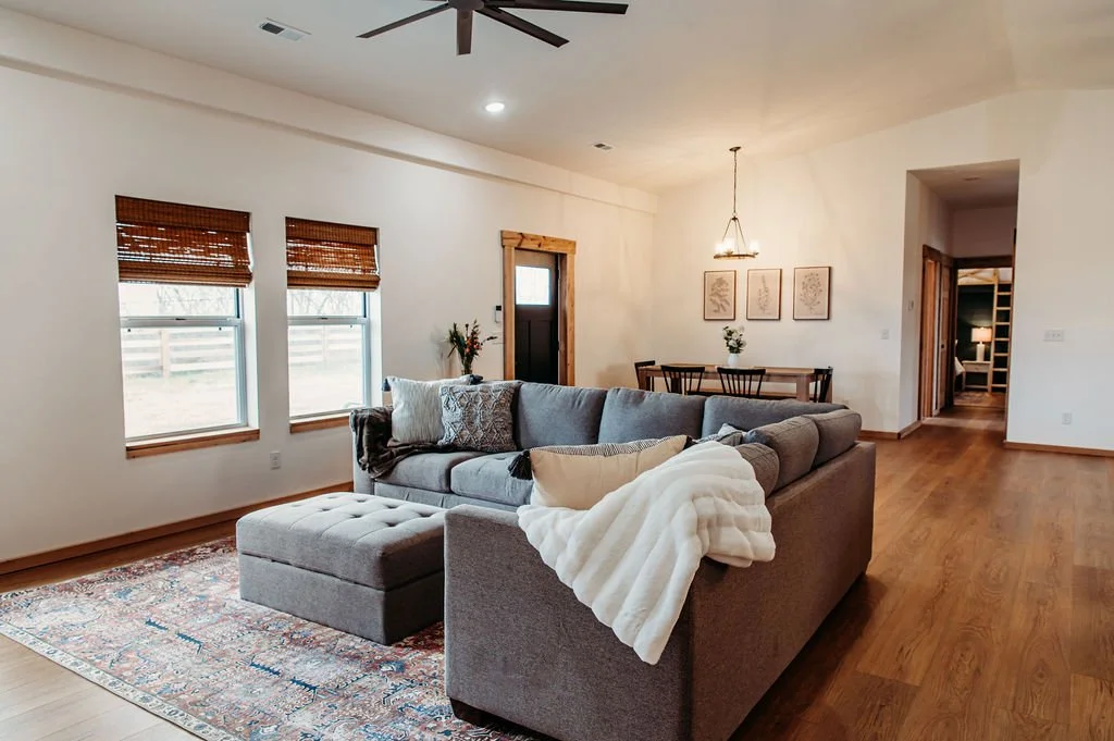 A living room with wooden floors, a gray sectional sofa with cushions and a throw blanket, three windows with bamboo blinds, and a dining table with chairs in the background. There is a chandelier and framed artwork on the wall.