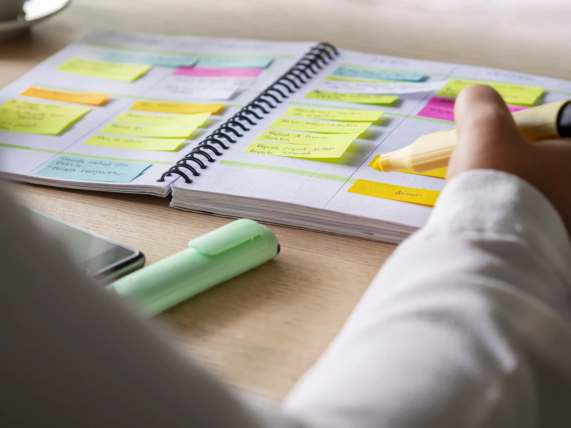Open planner with colorful sticky notes and a person writing in it, on a wooden desk. There is a green highlighter and a smartphone nearby.