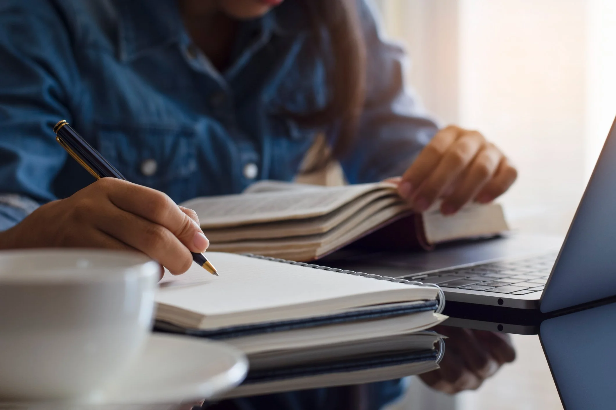 A person wearing a denim shirt writing in a notebook with a pen, with a laptop open nearby and stacked notebooks or papers on a desk.