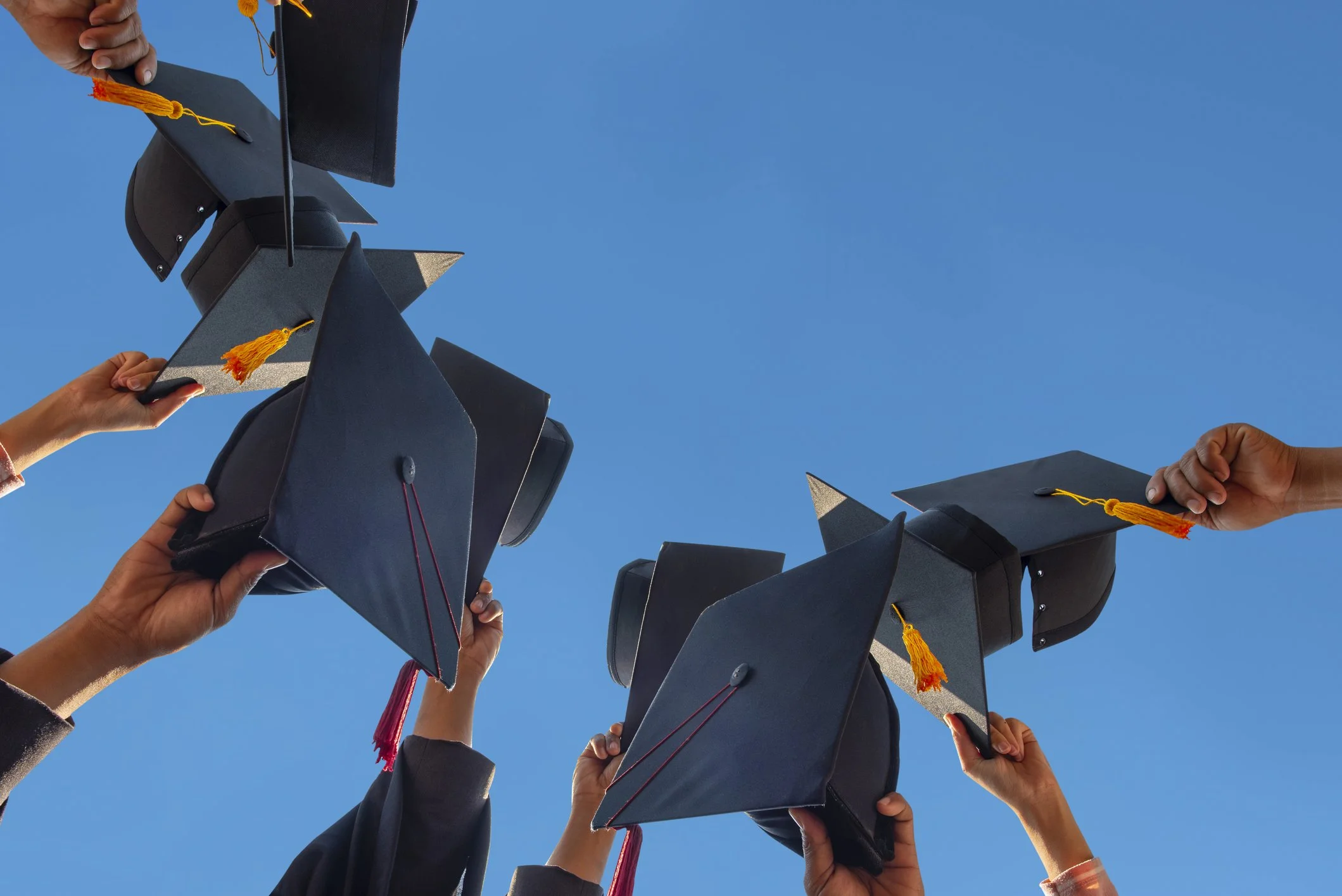 Several hands raising graduation caps against a clear blue sky.