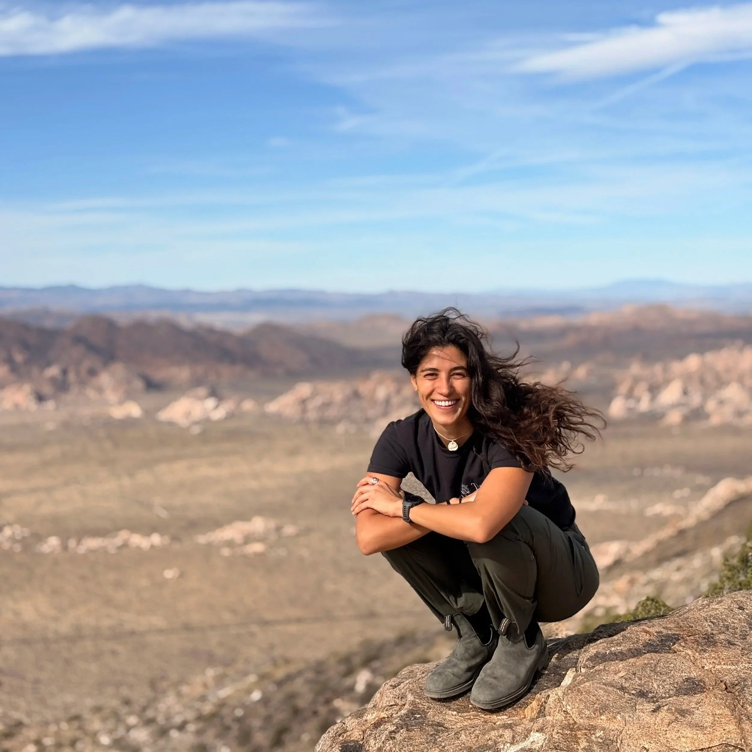 A smiling woman with curly hair, wearing a black shirt and a necklace, sitting on a tree log in a desert landscape with mountains and a blue sky with clouds.