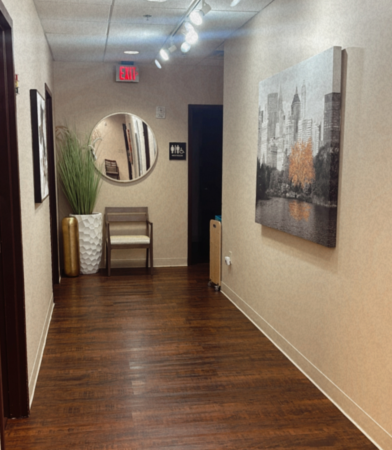 Empty hallway with wooden floor, beige walls, and modern artwork. Contains a large potted plant, a chair, and a mirror reflecting a chair inside a room. There is a sign indicating restrooms and a small heater with a white plug socket.