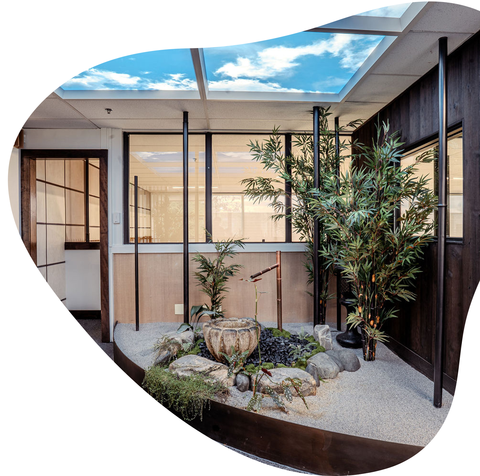 Indoor Japanese garden with a skylight, tall green plants, rocks, and a wooden and glass partition.