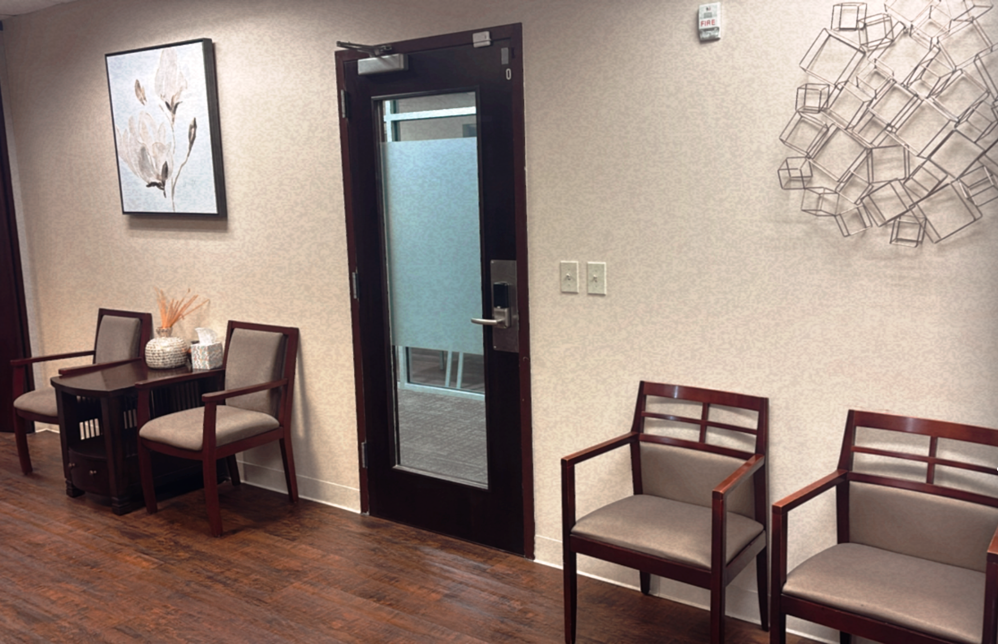 Waiting room with wooden chairs, a framed floral painting, and decorative wall art, near a door with frosted glass.