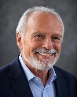 Close-up portrait of a smiling elderly man with white hair and beard, wearing a blue blazer and light blue shirt against a gray background.