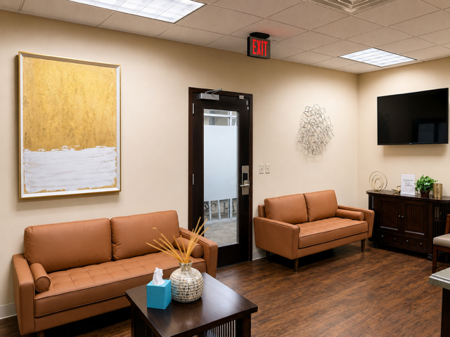 Waiting room with two brown sofas, a wooden coffee table with a vase and tissue box, wall art, a large TV, and an exit door, in an office or clinic setting.
