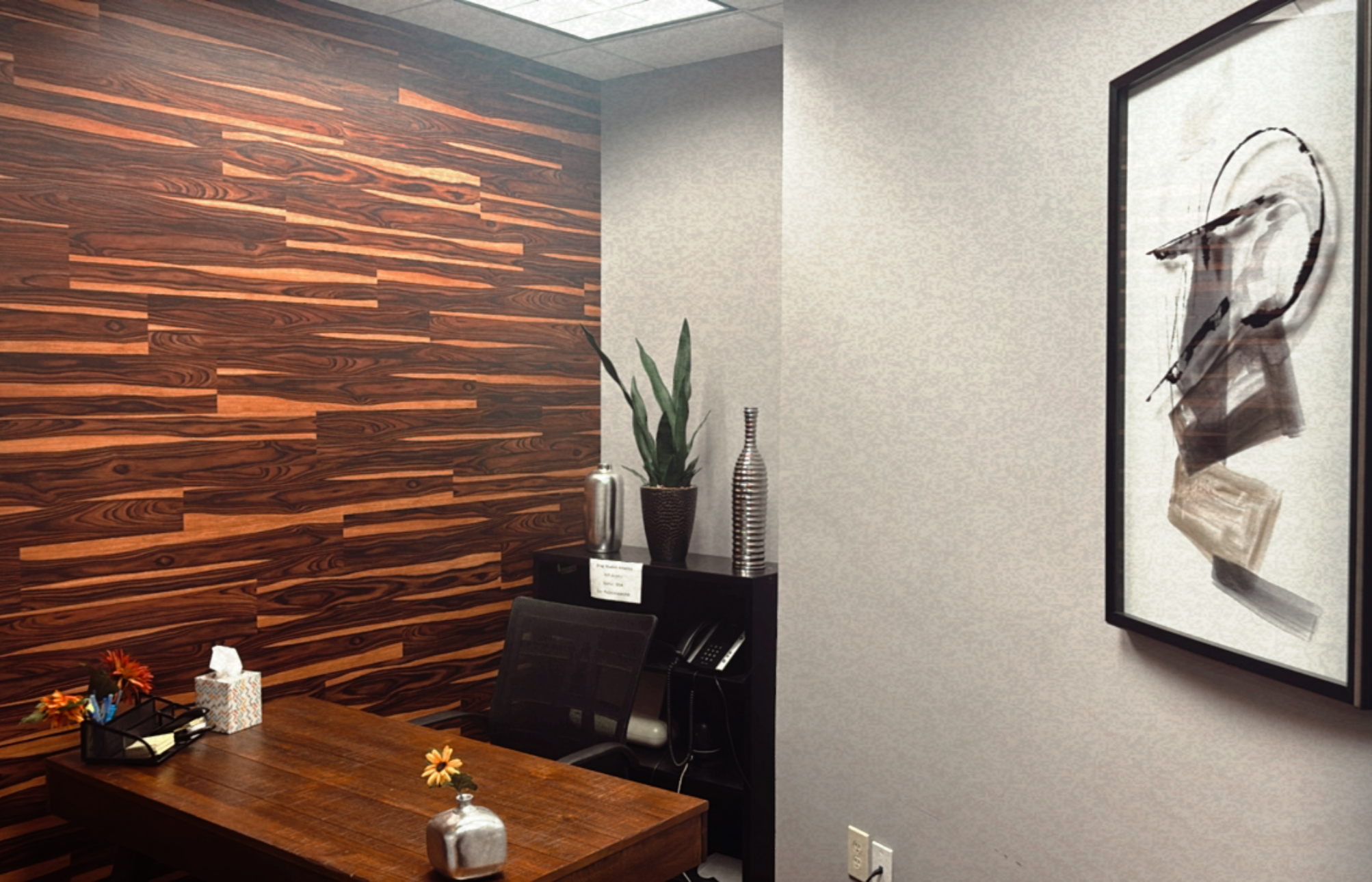 Office meeting room with a wood paneled wall, a desk with office supplies, a black chair, and framed abstract artwork on the wall.